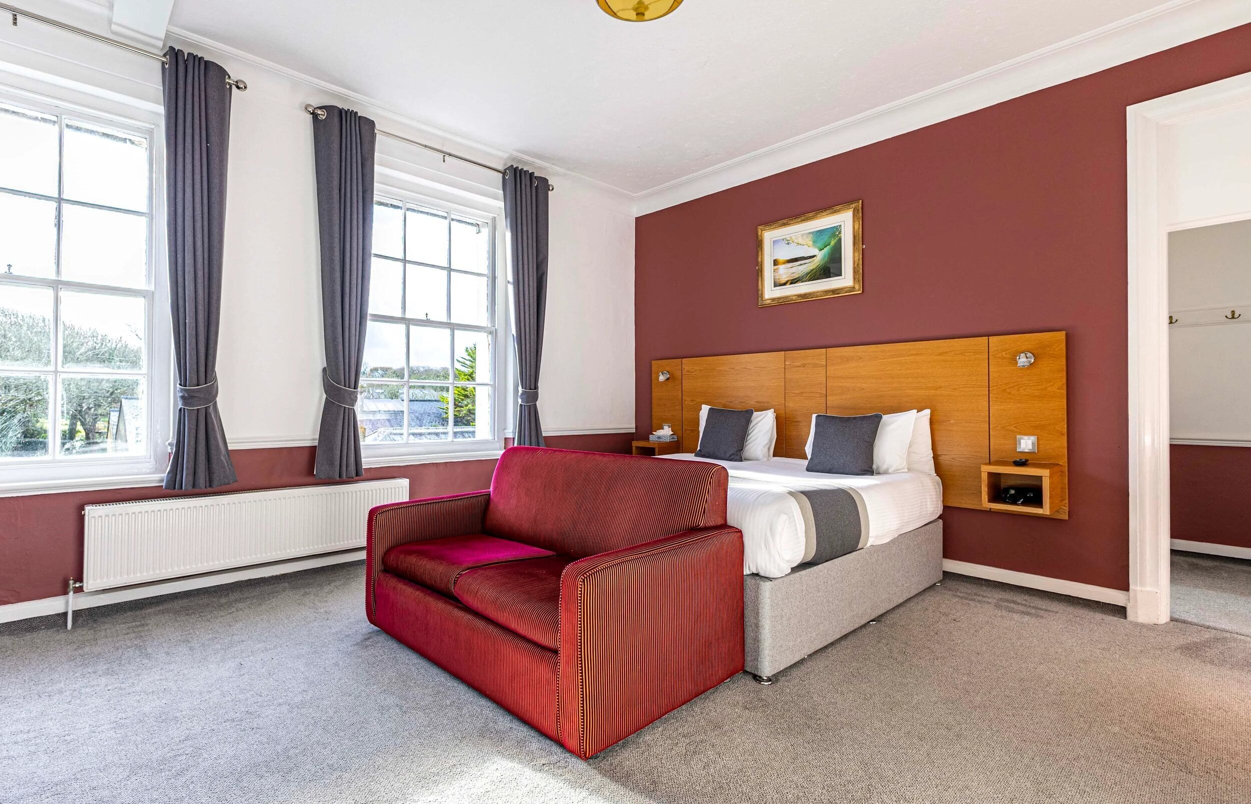 Habitación de hotel con cama, sofá rojo, cabecero de madera, ventana con cortinas y radiador.