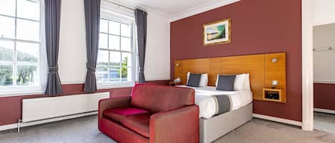 A hotel room with a bed, a red sofa, a wooden headboard, a window with curtains, and a radiator.