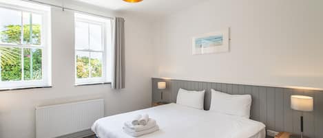 Habitación de hotel ordenada, con una cama grande, dos mesitas de noche con lámparas, una ventana con cortinas, un radiador y un cuadro en la pared.