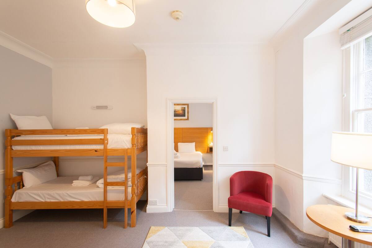 A hotel room with a bunk bed, a single bed, a red chair, and a small table with a lamp.