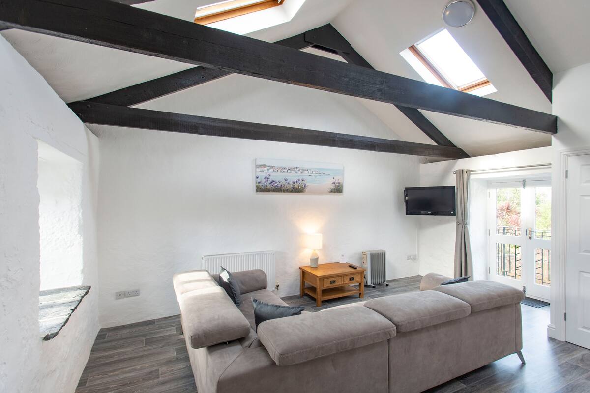 A modern living room with a high ceiling, exposed beams, a large window, a sofa, a wooden coffee table, and a television.