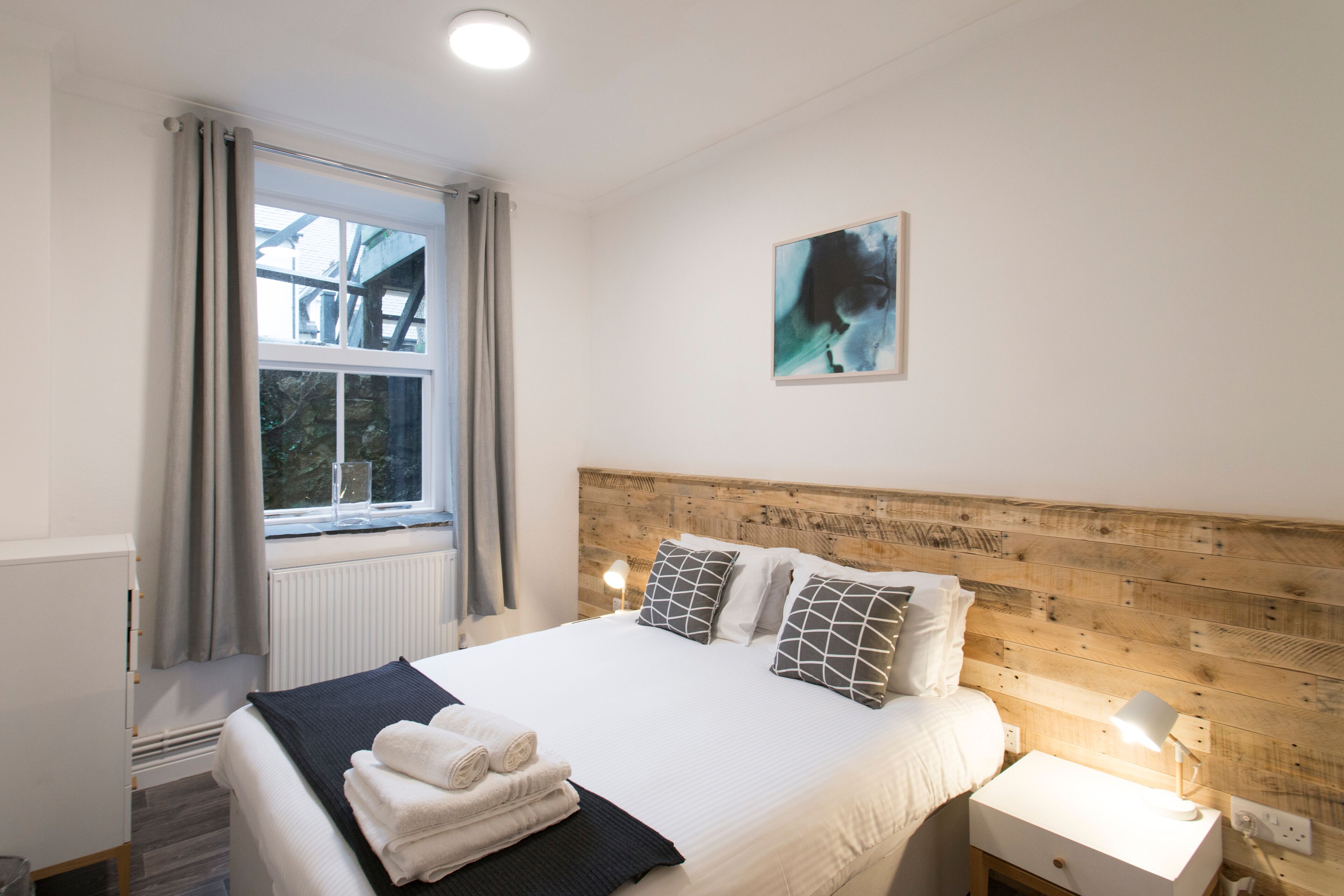 A neatly made bed with a wooden headboard, a bedside table with a lamp, and a window with curtains.