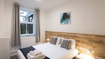 Una cama bien hecha con cabecera de madera, una mesita de noche con lámpara y una ventana con cortinas.
