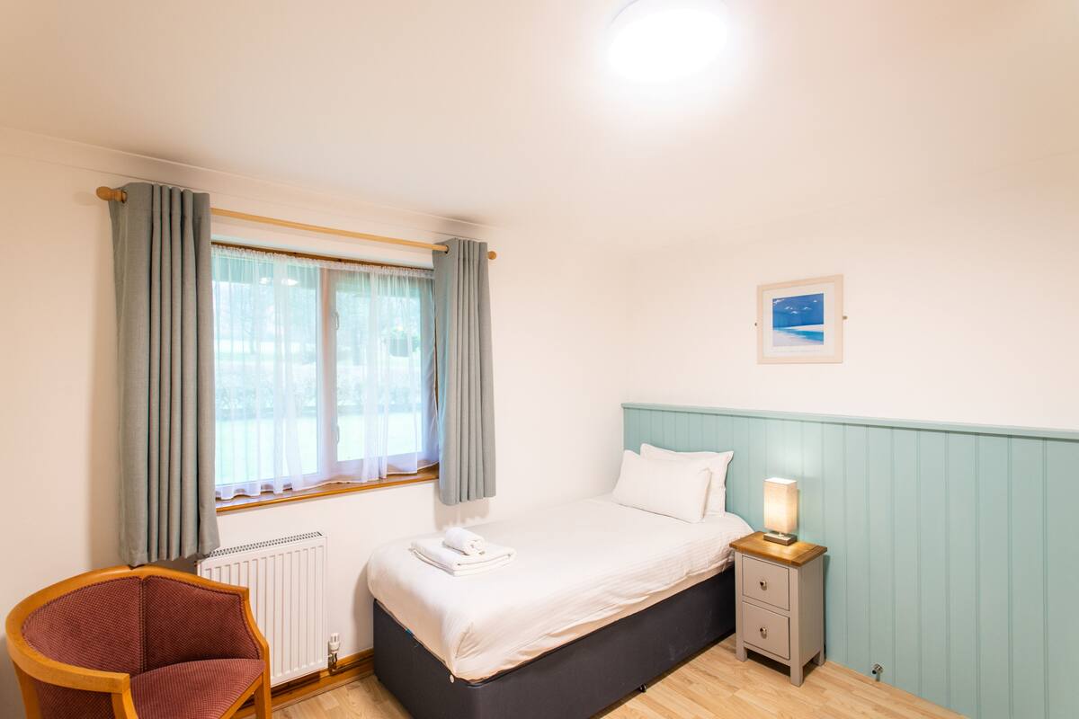A bedroom with a bed, a nightstand, a chair, a window with curtains, and a picture on the wall.