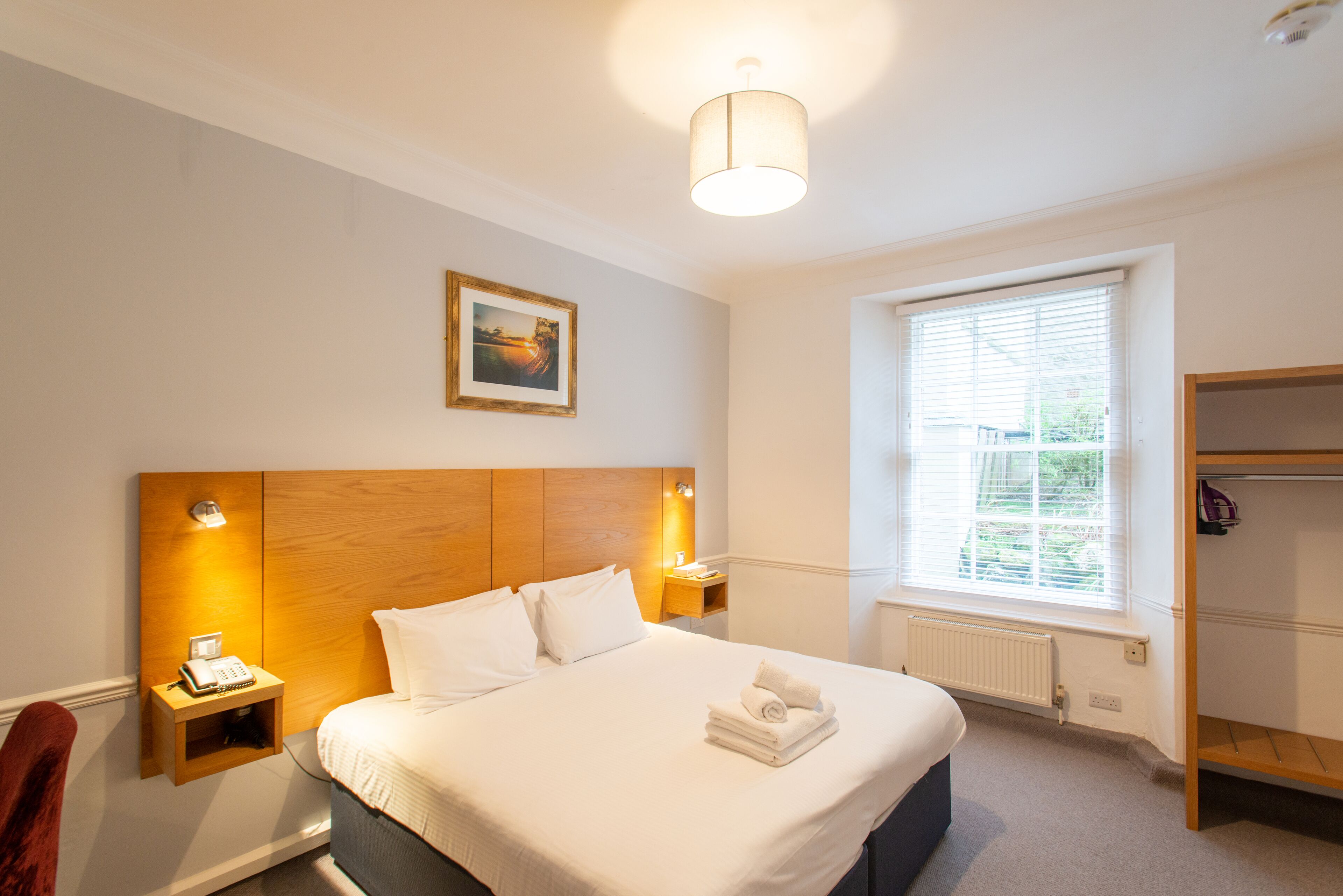 A hotel room with a large bed, bedside lamps, a framed picture on the wall, a window with blinds, and a wooden wardrobe.