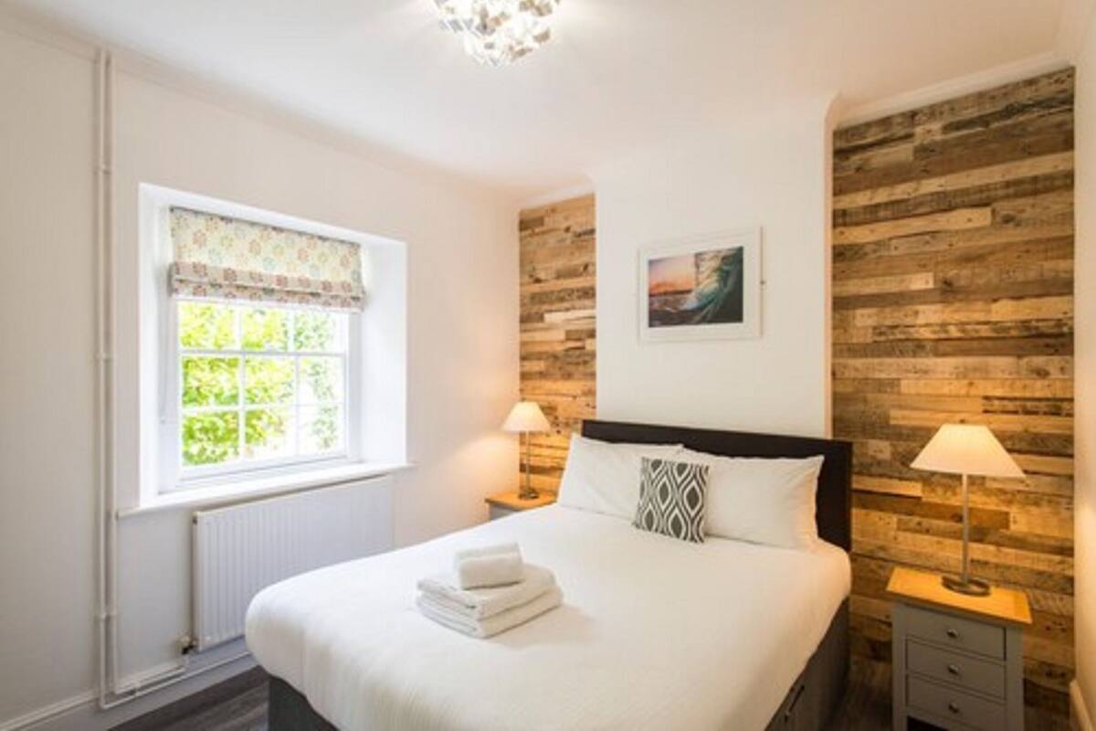 A bedroom with a large bed, two bedside tables with lamps, a window with a floral valance, and a wooden wall panel.