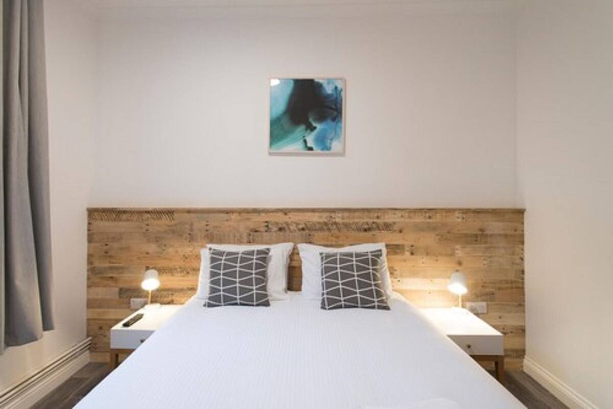 A bedroom with a wooden headboard, a bed with white bedding and geometric pillows, two bedside tables with lamps, and a framed picture on the wall.