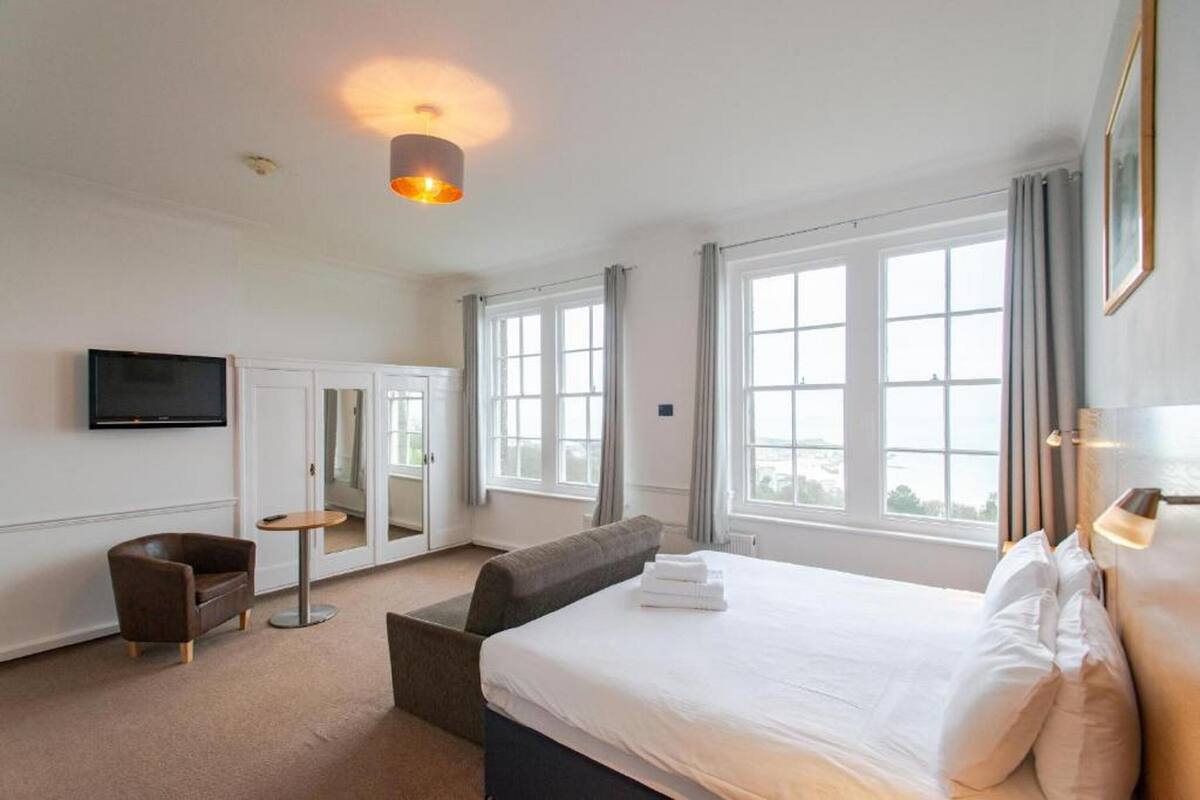 A hotel room with a large bed, a sofa, a small table, a TV, and large windows.