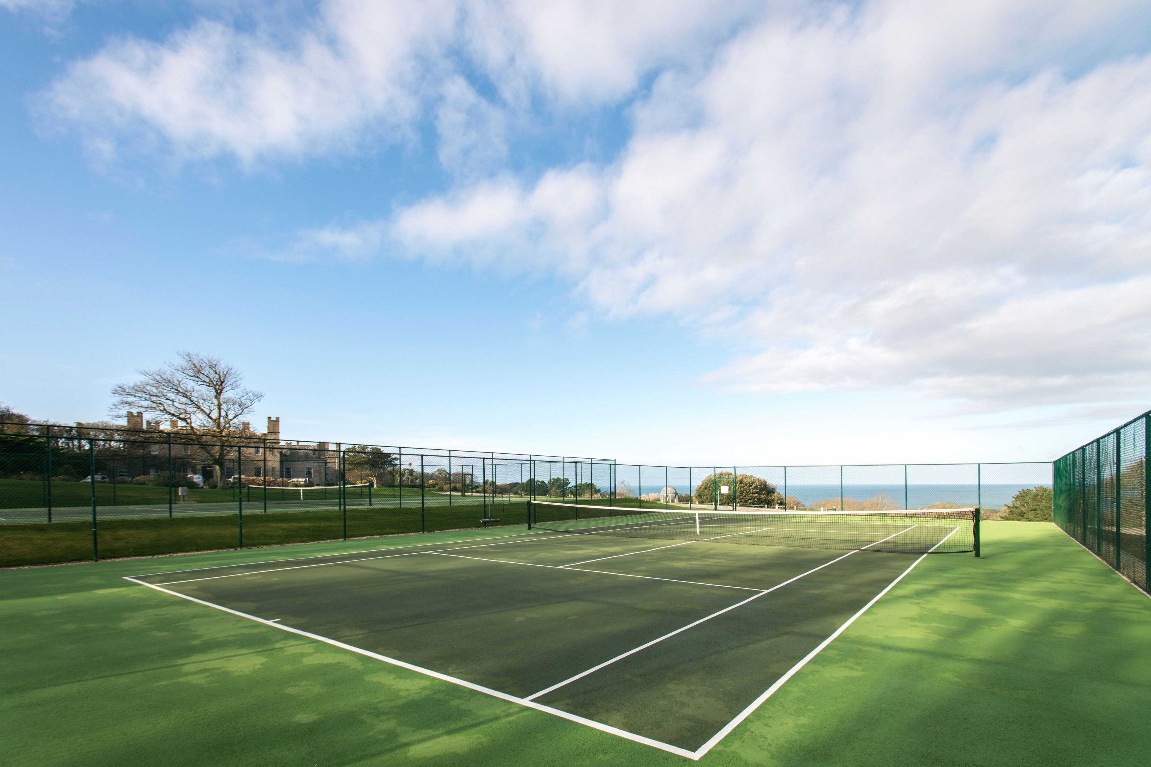 A tennis court with a net, surrounded by a green fence, and a clear sky with clouds.