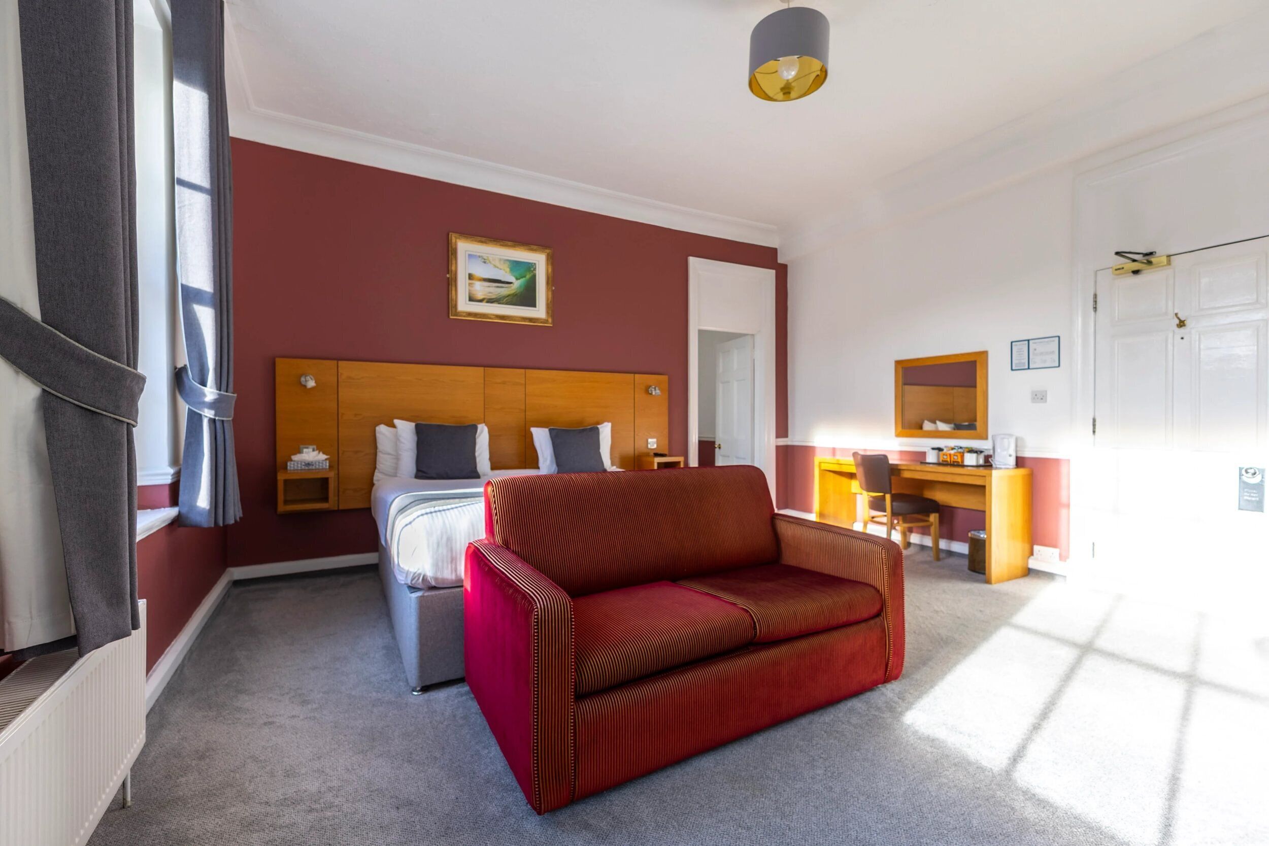 Quarto de hotel com cama, sofá vermelho, escrivaninha e janela.