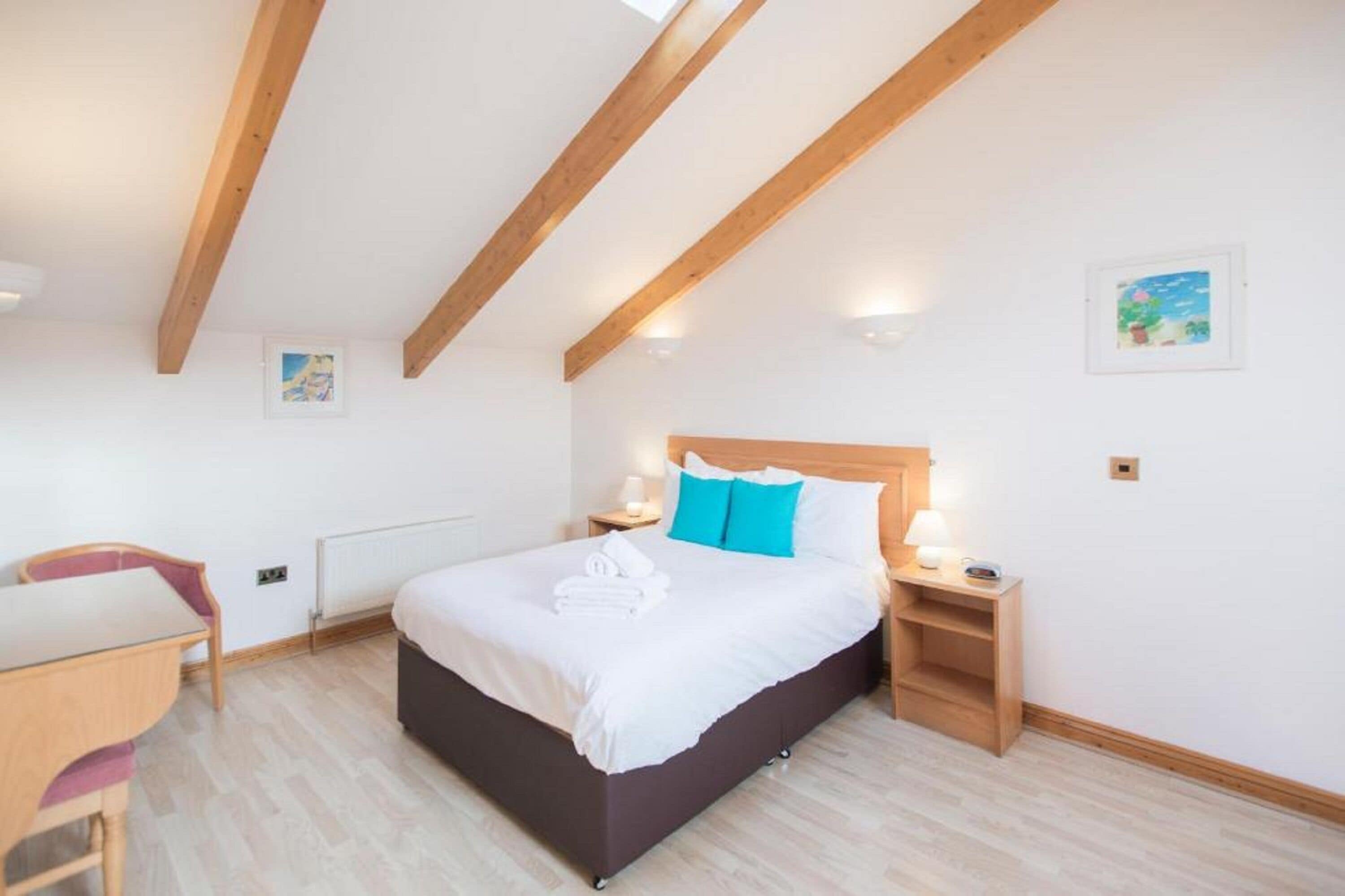A bedroom with a wooden ceiling, a bed with white bedding and blue pillows, a bedside table with a lamp, and a small wooden desk with a chair.