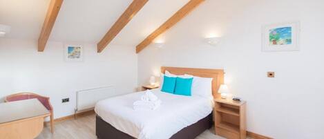 A bedroom with a wooden ceiling, a bed with white bedding and blue pillows, a bedside table with a lamp, and a small wooden desk with a chair.