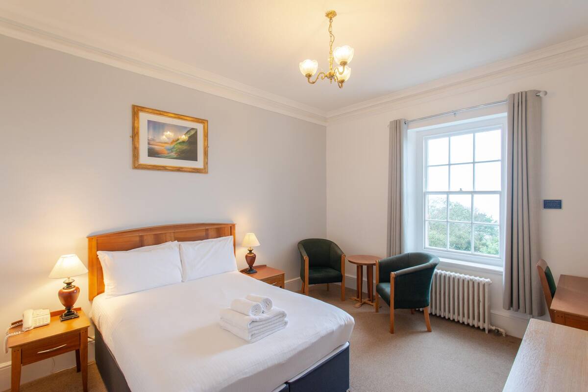 A hotel room with a large bed, a bedside table with a lamp, a chair, a small table, and a window with curtains.