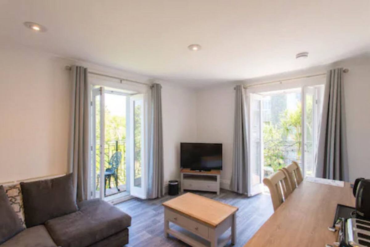 A modern living room with a sofa, a coffee table, a TV on a stand, and a dining table with chairs. There are large windows with curtains, and a balcony is visible through the windows.