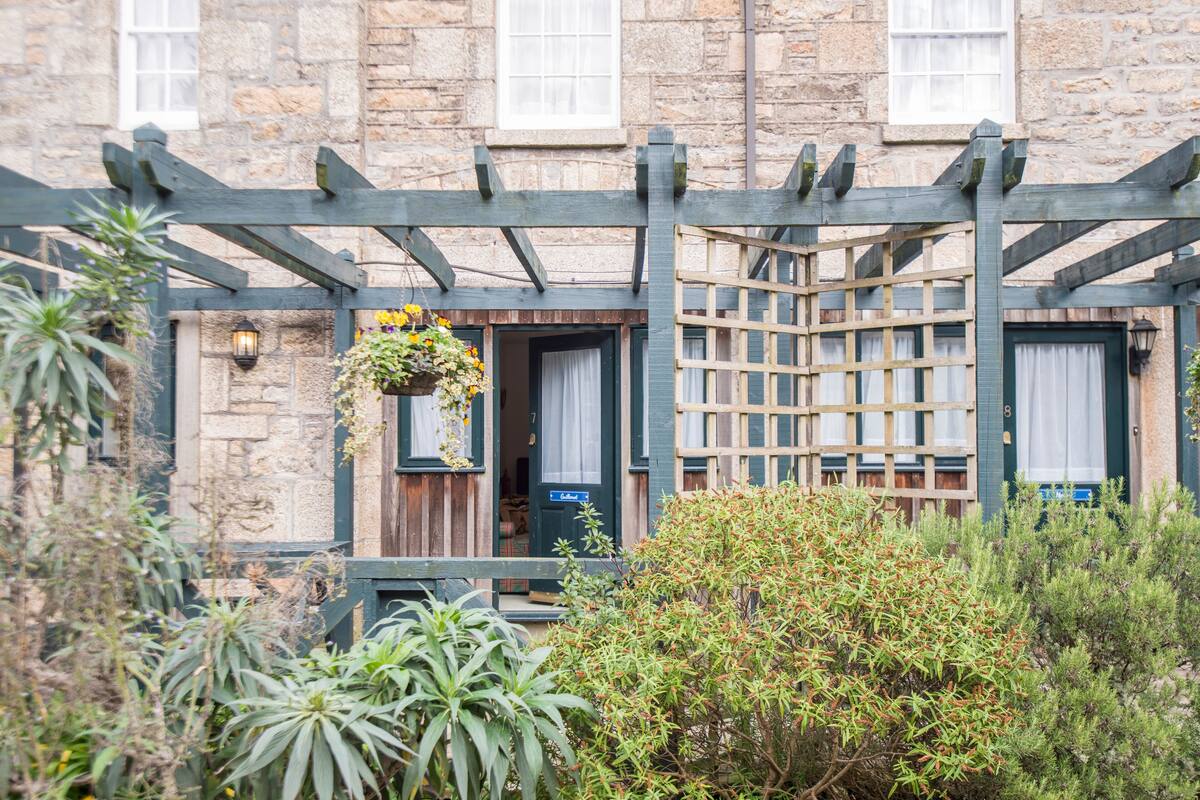 A stone building with a wooden pergola, a hanging flower basket, and a door with a sign.