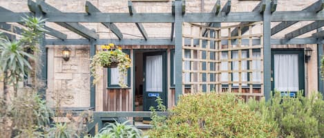 A stone building with a wooden pergola, a hanging flower basket, and a door with a sign.