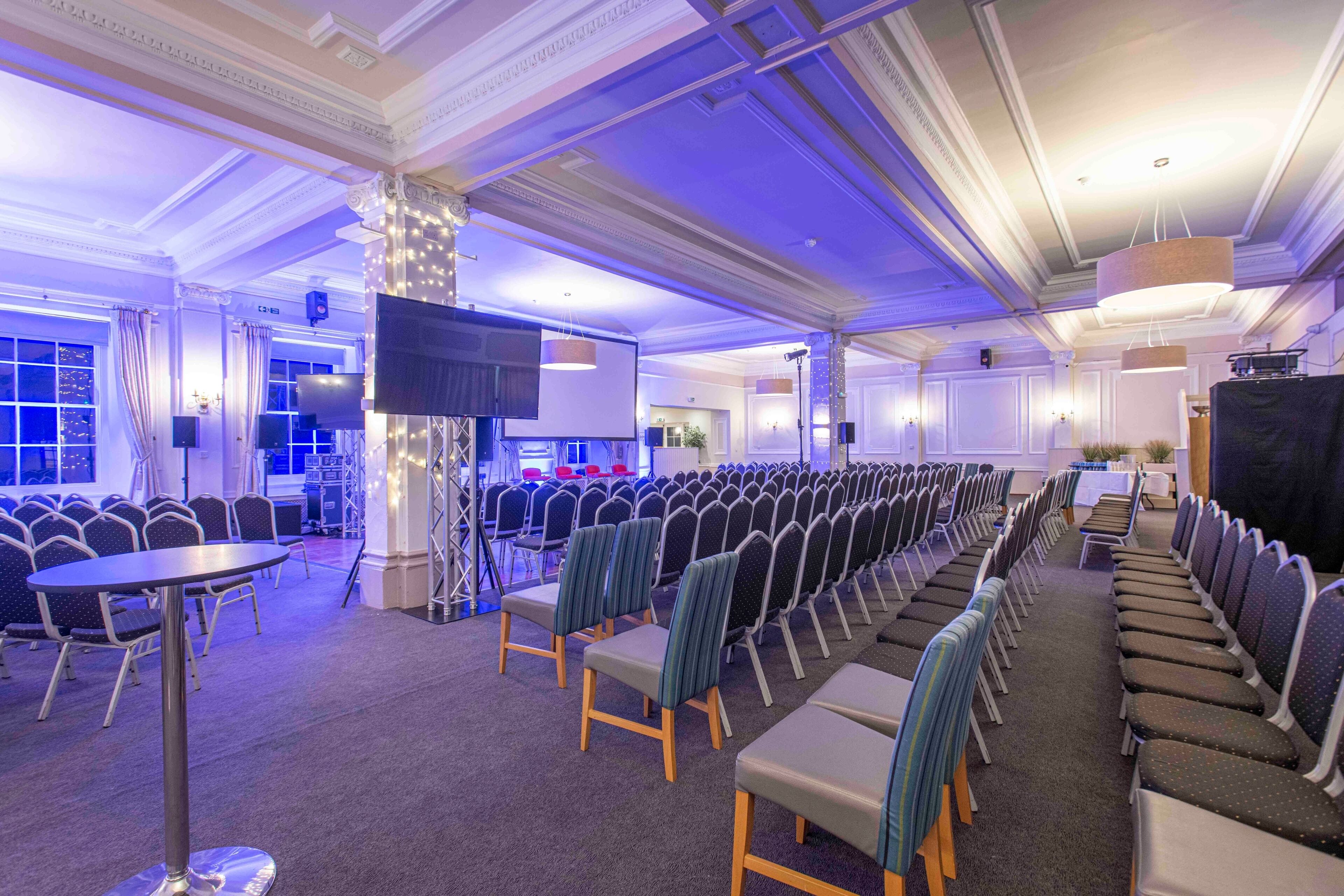 A conference room with rows of chairs, a stage with a screen, and a large pillar with lights.