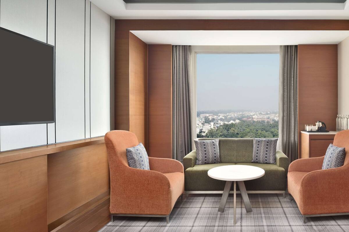 Une chambre d’hôtel moderne avec vue sur la ville, deux fauteuils, une petite table ronde et un canapé agrémenté de coussins à motifs.