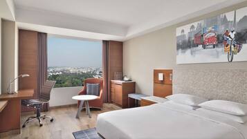 A hotel room with a large bed, a desk, a chair, a small table, and a view of a cityscape.