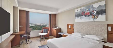 A hotel room with a large bed, a desk, a chair, a small table, and a view of a cityscape.