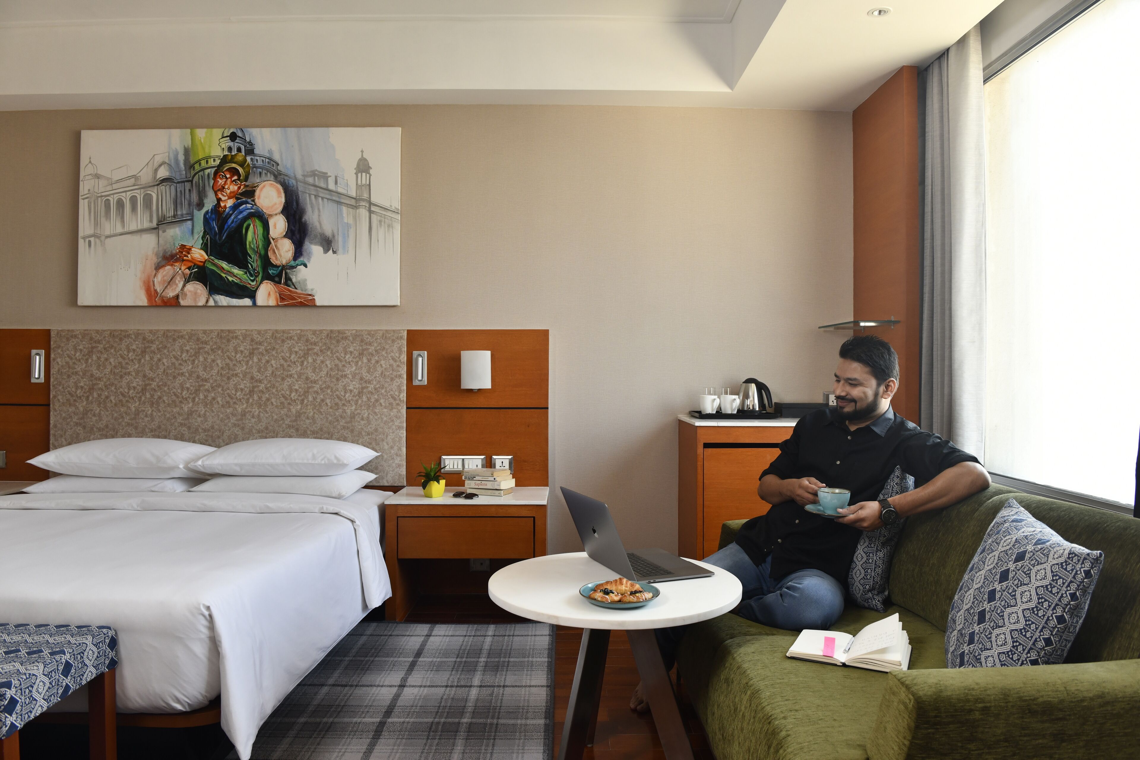A hotel room with a bed, a desk with a laptop, a dining table with a plate of food, and a painting on the wall.