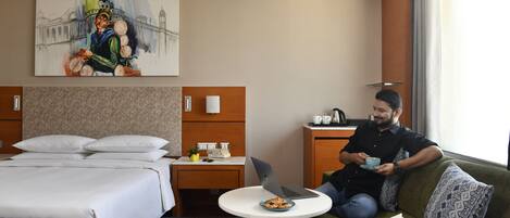 A hotel room with a bed, a desk with a laptop, a dining table with a plate of food, and a painting on the wall.