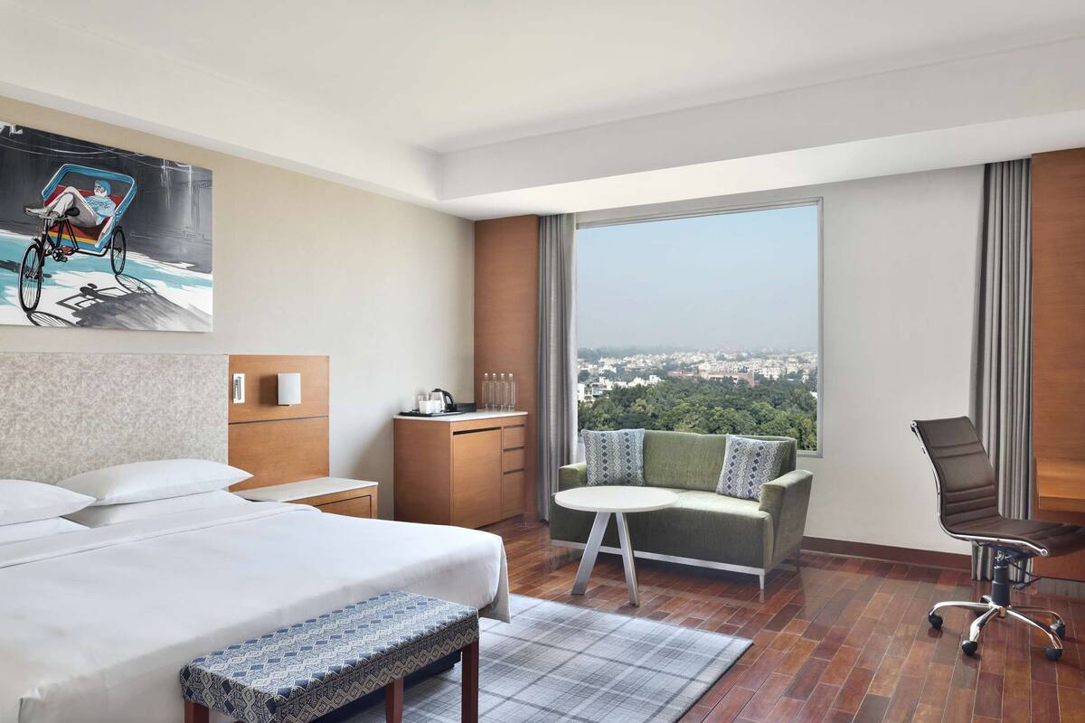 Une chambre d’hôtel avec un grand lit, un bureau, une chaise, un banc, une grande fenêtre donnant sur la ville et un tableau au mur.