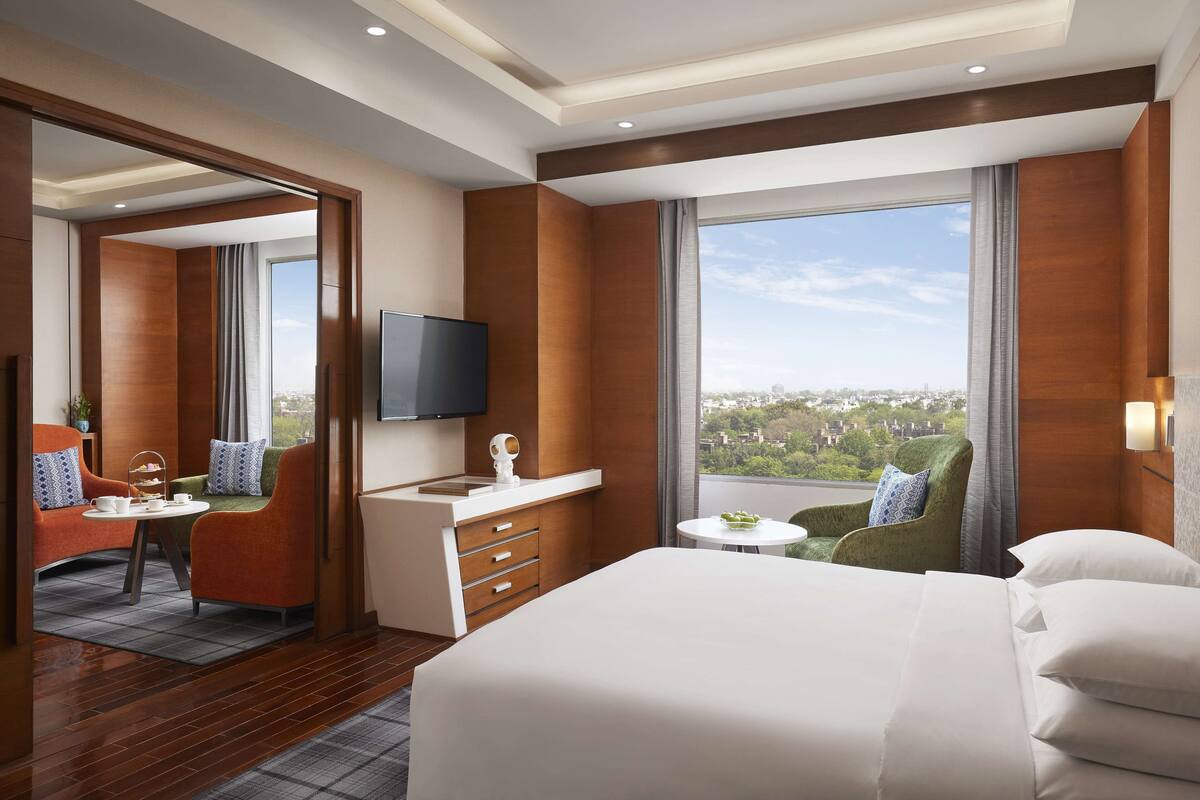 Une chambre d’hôtel dotée d’un grand lit, d’un bureau avec un téléviseur à écran plat, d’un coin salon avec des fauteuils et offrant une vue sur la ville grâce à de grandes fenêtres.