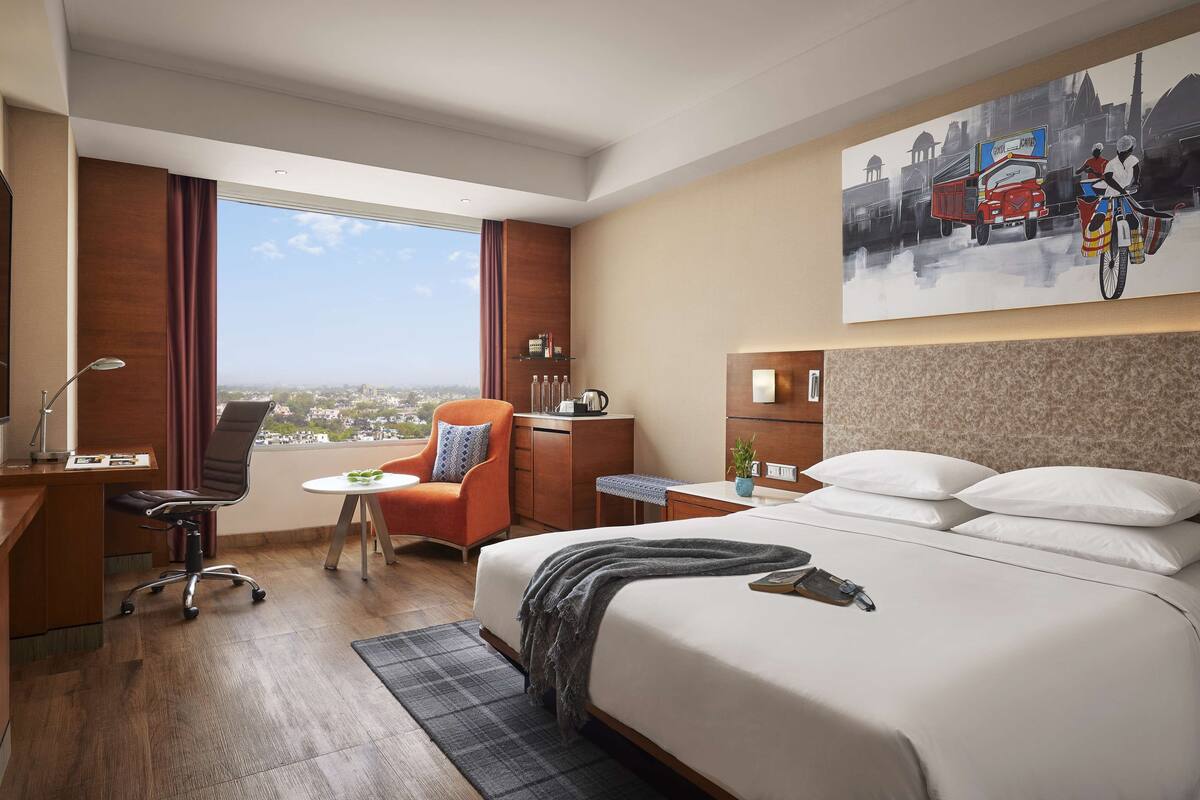 Une chambre d’hôtel avec un grand lit, un bureau, une chaise, une petite table, une table de chevet, une lampe, un tableau et une vue sur la ville.