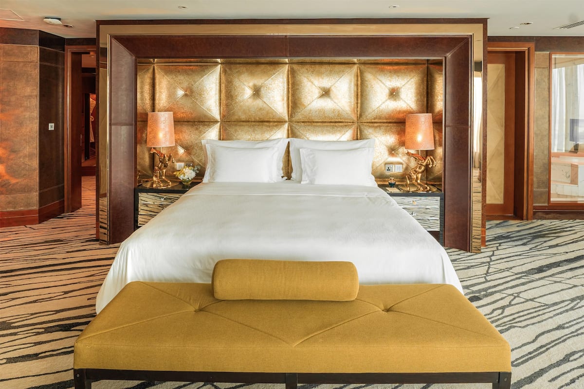 A hotel room with a large bed, a gold-toned headboard, two bedside lamps, and a patterned carpet.