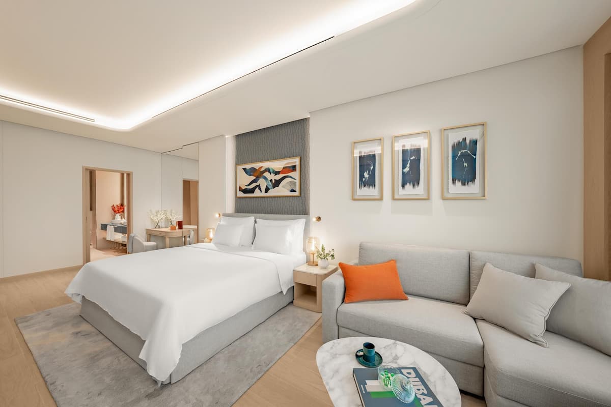 A modern hotel room with a large bed, a grey sofa, and a small table with a cup and a book. There are three framed pictures on the wall and a mirror above the bed.