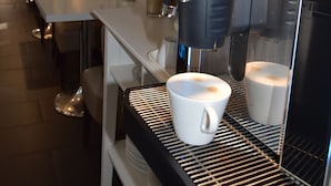 Uma máquina de café com menu digital, uma chávena branca no balcão e um vaso com flores vermelhas.