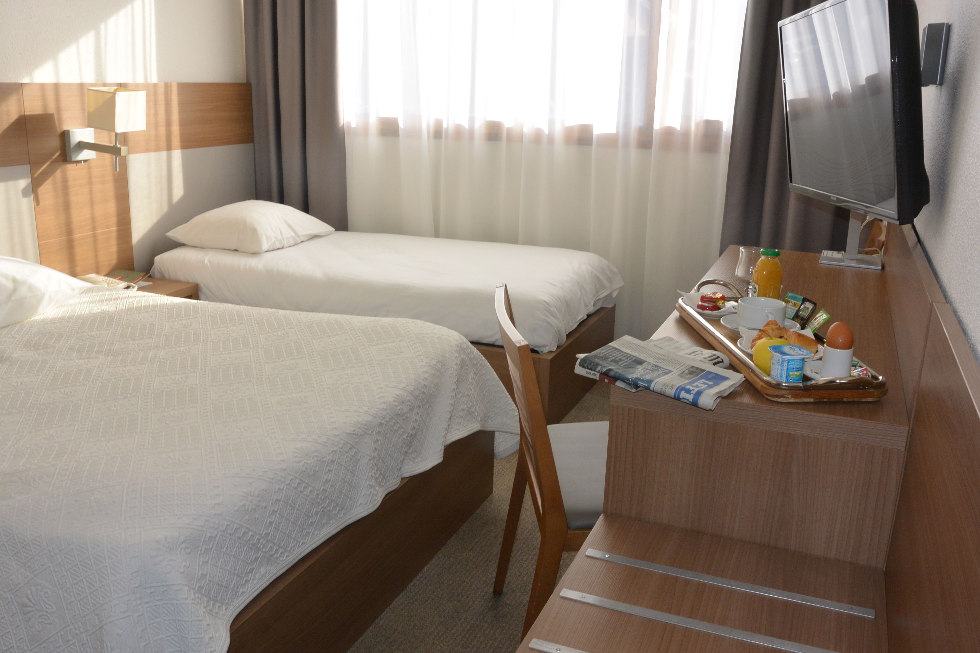 A hotel room with two beds, a desk with breakfast items, a TV, and a window with curtains.