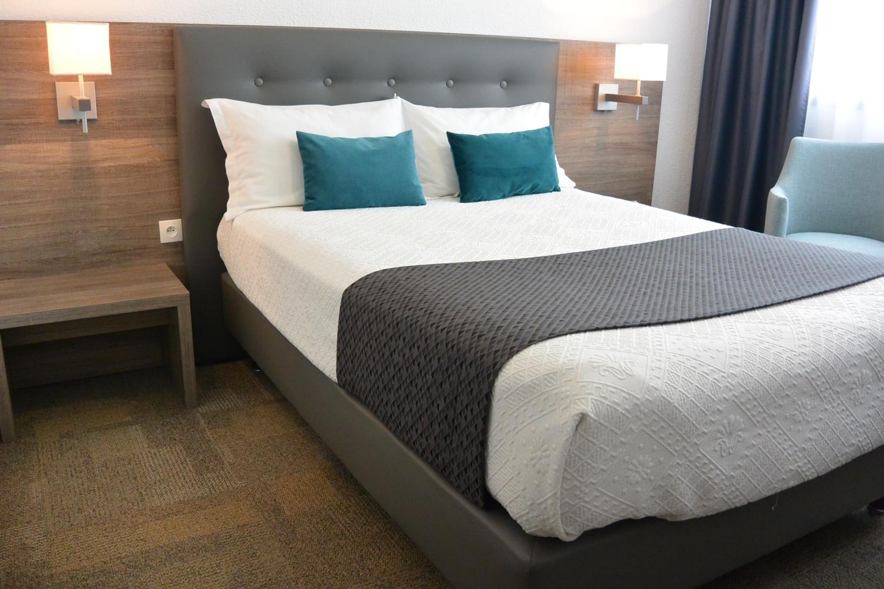 A neatly made bed with a grey headboard, two white pillows, and two teal pillows. There is a grey throw blanket on the bed. The room has a wooden headboard, a nightstand with a lamp, and a chair.