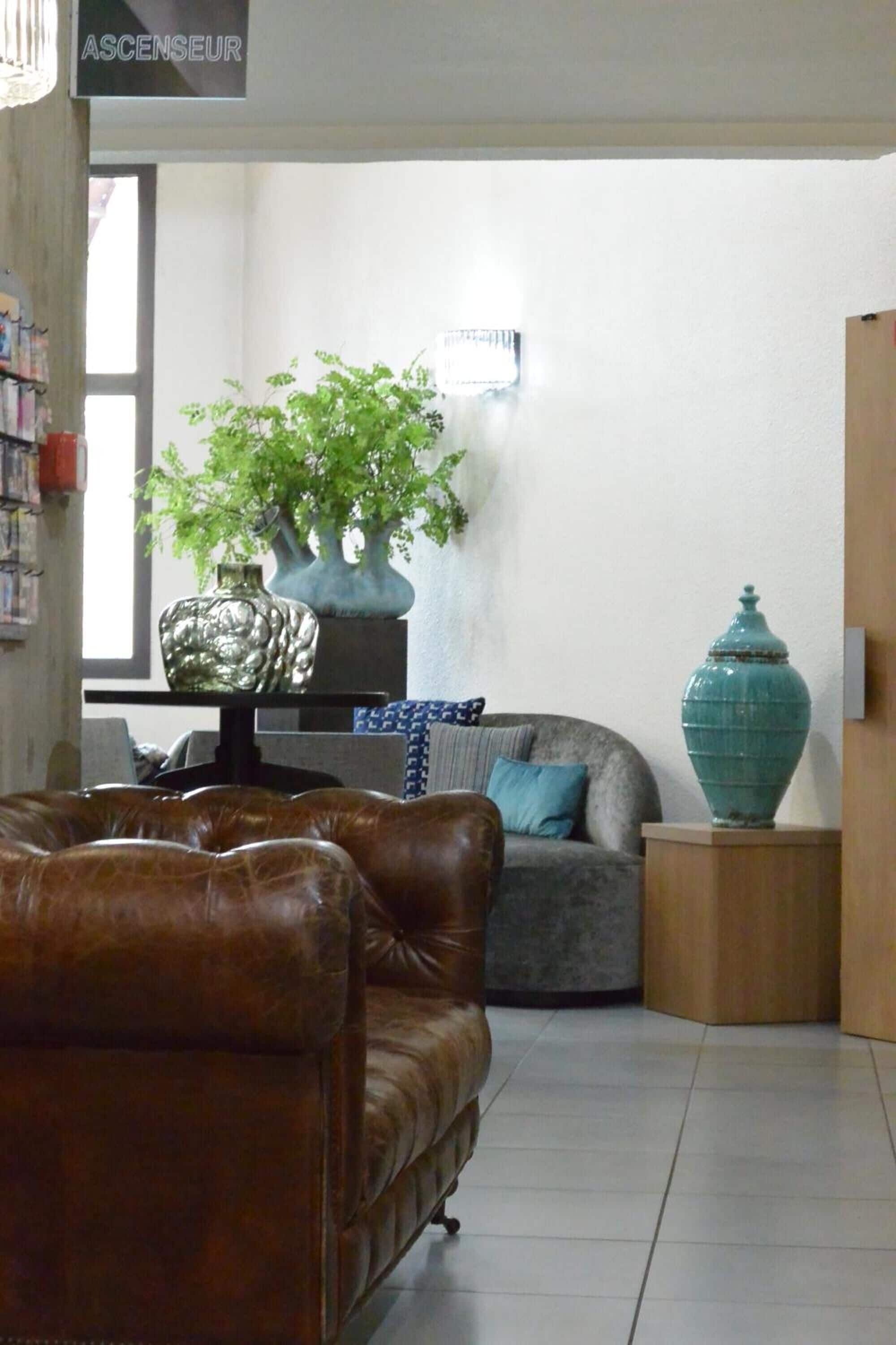 A living room with a brown leather sofa, a grey armchair, a large turquoise vase, and a potted plant on a table.