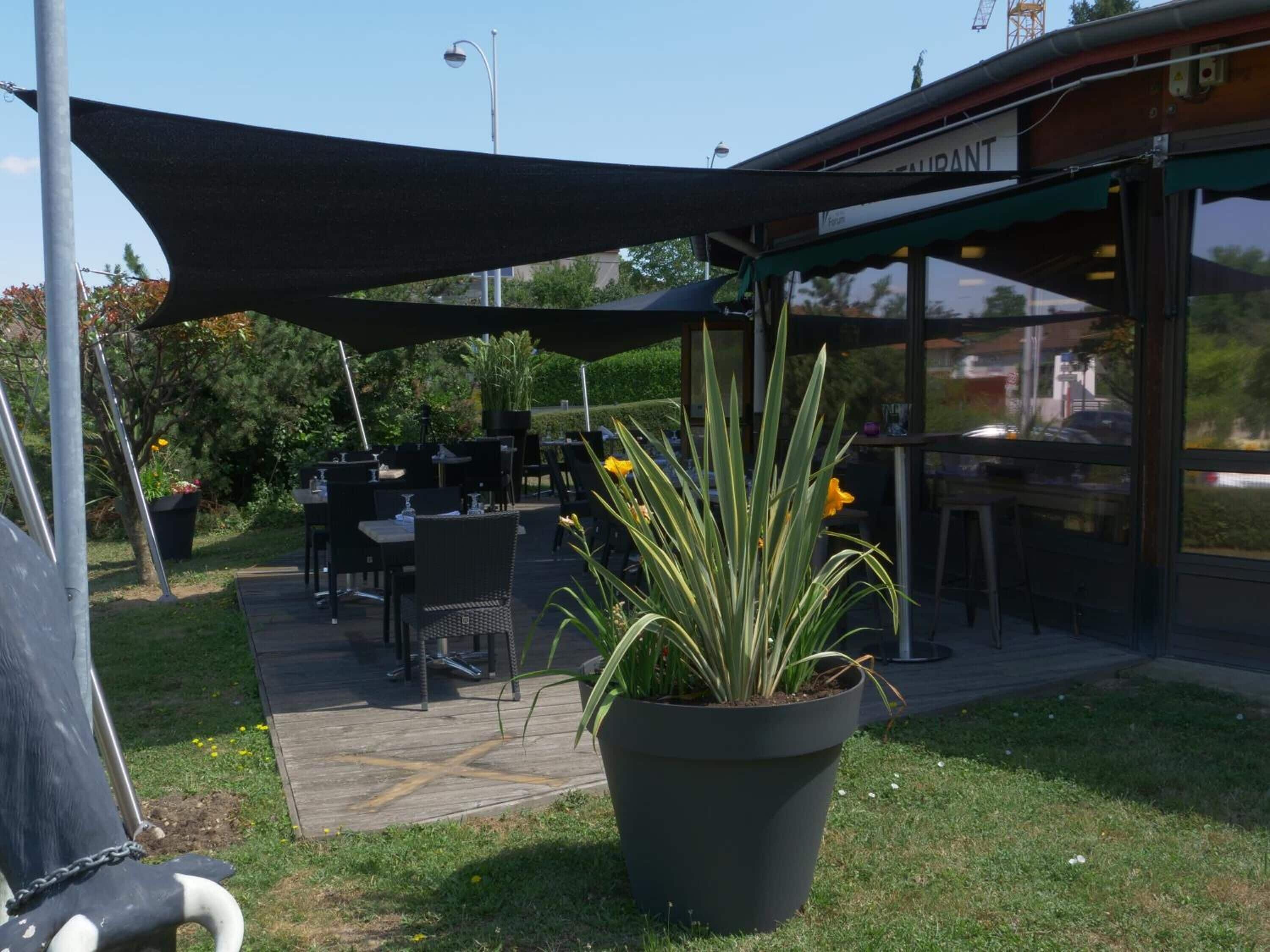 An outdoor seating area with tables and chairs under a shaded canopy, a potted plant, and a building with a sign that reads "SUPRANT".