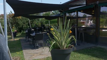 An outdoor seating area with tables and chairs under a shaded canopy, a potted plant, and a building with a sign that reads "SUPRANT".