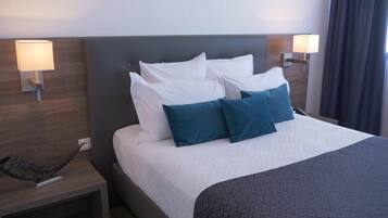 Cama bem arrumada com almofadas brancas e azul-esverdeadas, cabeceira escura e duas luminárias de cabeceira em mesas de cabeceira de madeira.