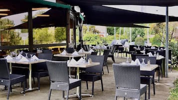 Een buiten eethoek met tafels en stoelen, beschaduwd door parasols en omgeven door groen.
