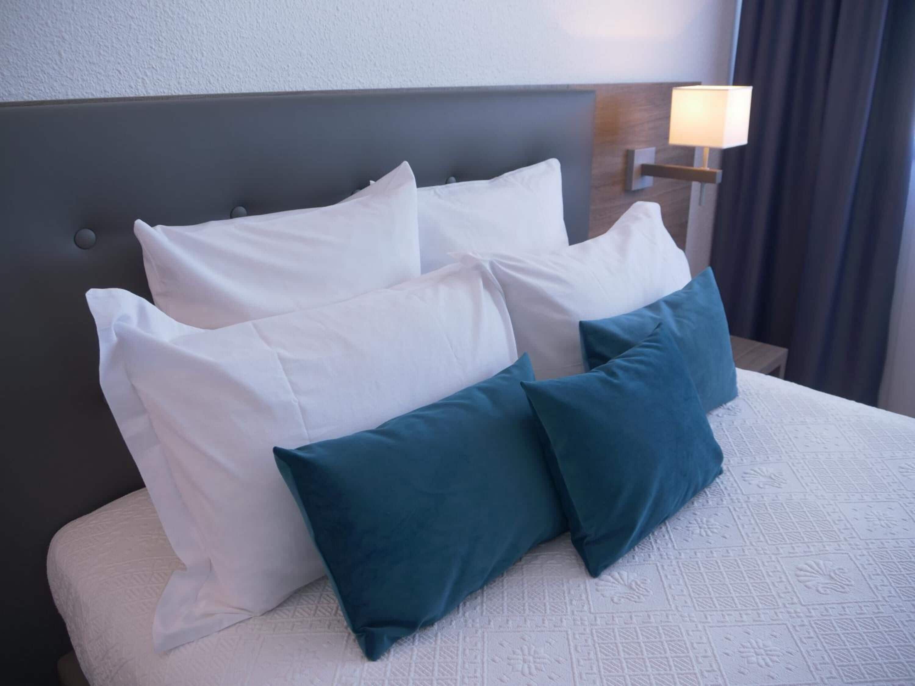 A bed with white and teal pillows, a textured white bedspread, and a wall-mounted lamp.