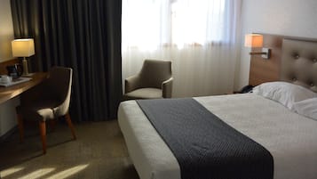 Une chambre d’hôtel avec un lit, un bureau avec une lampe, une chaise et une fenêtre avec des rideaux.