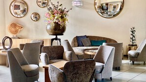 A modern lounge area with multiple seating options, including sofas and armchairs, arranged around a central coffee table. There are several wall-mounted mirrors and a decorative vase with flowers. - Kyriad Lyon Est - Genas Eurexpo (Genas)