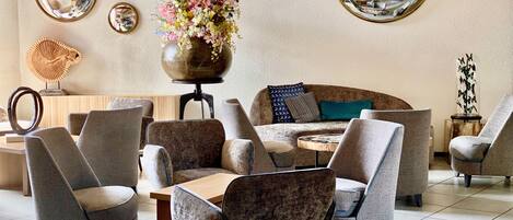 Un moderno salón con varias opciones de asientos, incluyendo sofás y sillones, dispuestos alrededor de una mesa de centro. Hay varios espejos montados en la pared y un jarrón decorativo con flores.