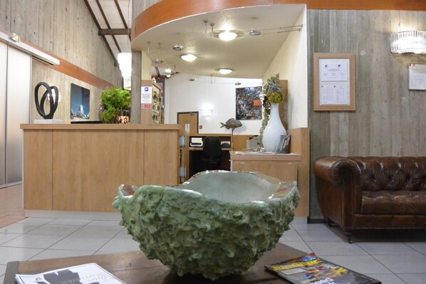 A reception area with a wooden counter, a green ceramic bowl, a brown leather couch, and a wall with framed documents. - Kyriad Lyon Est - Genas Eurexpo (Genas)