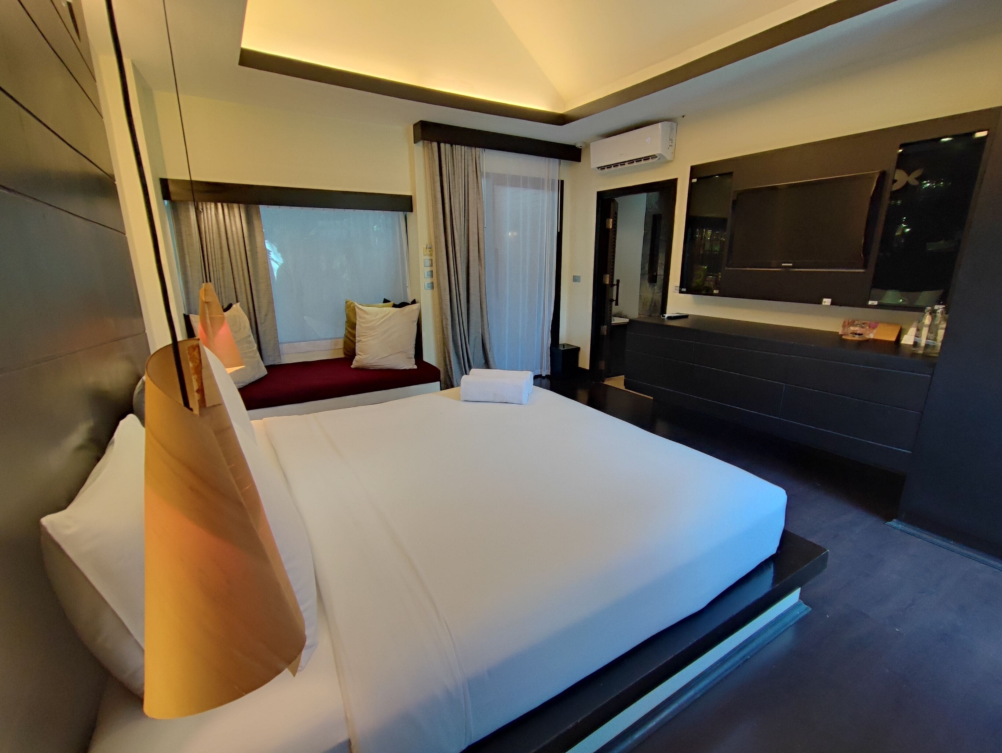 A modern hotel room with a large bed, a flat-screen TV, a minibar, and a large window with curtains.
