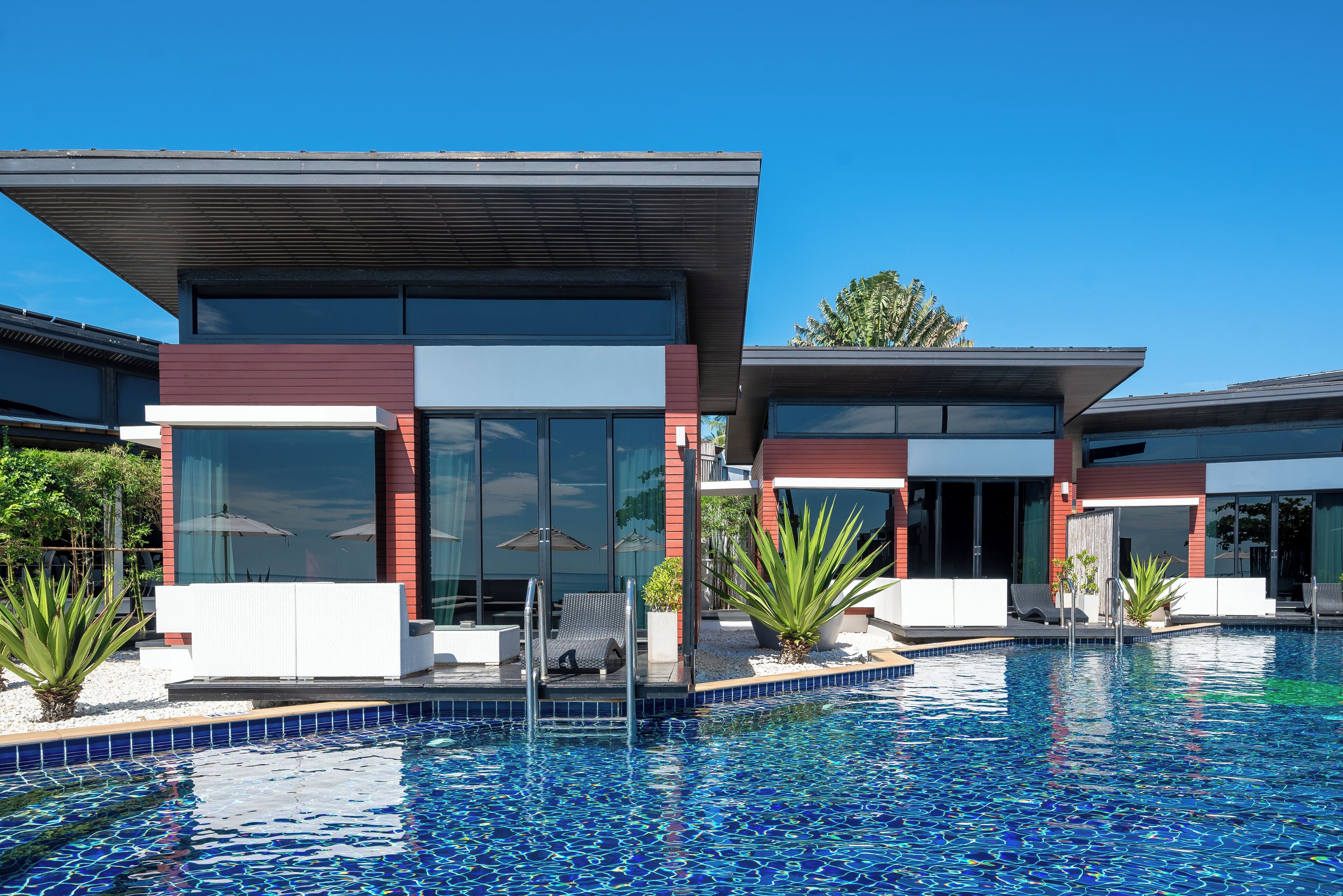 Un espace moderne aménagé au bord de la piscine, avec une piscine à l’eau d’un bleu limpide, des chaises longues et un bâtiment contemporain doté de grandes fenêtres et d’une façade rouge.