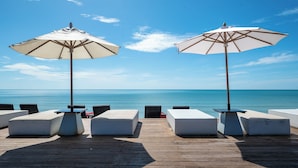Eine Holzterrasse mit weiß gepolsterten Sitzmöbeln und großen Sonnenschirmen, mit Blick auf ein ruhiges Meer.