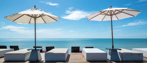 A wooden deck with white cushioned seating and large umbrellas, overlooking a calm sea.