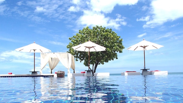 Ein Pool ohne Ende mit klarem, blauem Wasser, umgeben von weißen Liegestühlen und Sonnenschirmen, und ein Baum in der Mitte.