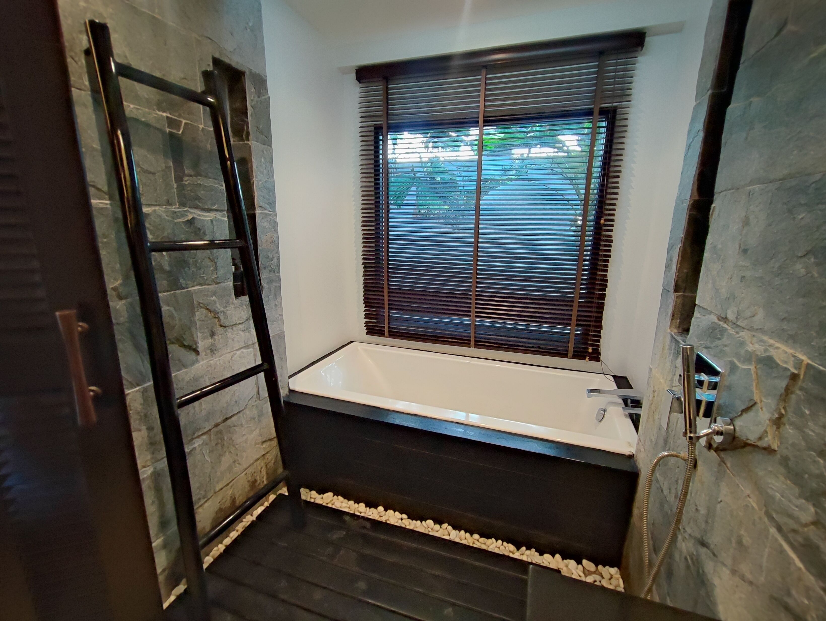 A bathroom with a bathtub, stone walls, a window with blinds, and a towel rack.